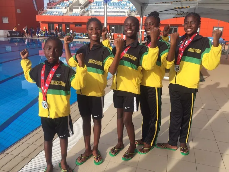 Jamaican swimmers in green and gold team kit holding their medals beside the pool